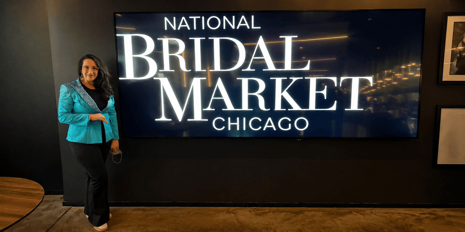 Person in a teal blazer poses beside a large screen displaying "National Bridal Market Chicago"