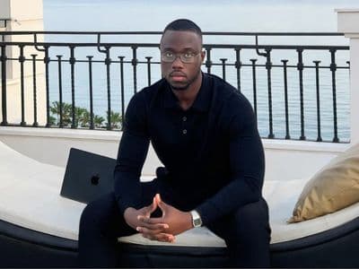 A man in a black polo shirt sits outdoors by the ocean with an Apple laptop, reflecting on sales systems.