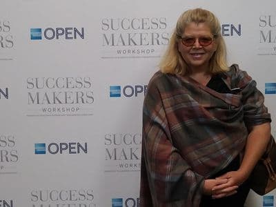 Janet Walker posing at Success Makers Workshop event backdrop wearing plaid shawl and sunglasses.
