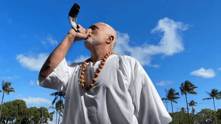 Man dressed in white, blowing a horn under a bright sky, symbolizing spiritual outreach and unity.