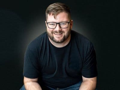 Will Craig smiling in glasses and a black shirt against a dark background.