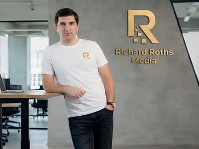 A person in a white shirt with a logo leans on a table in a modern office. The wall displays "Richard Roths Media" in gold. The atmosphere is professional.
