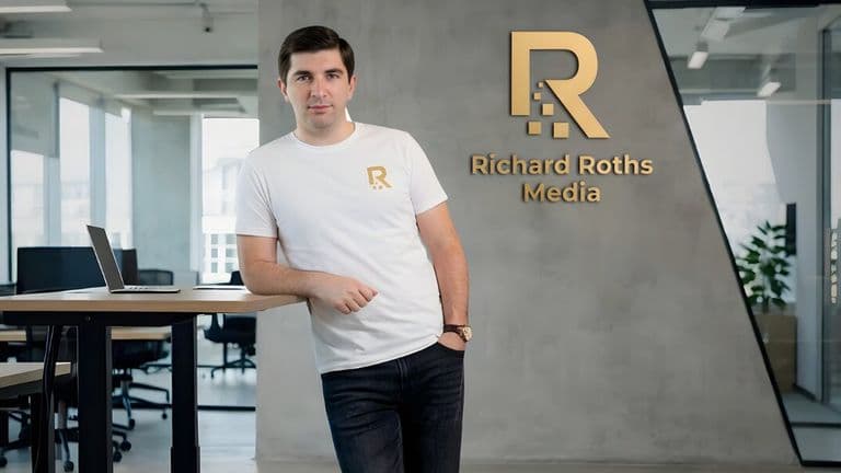 A person in a white shirt with a logo leans on a table in a modern office. The wall displays "Richard Roths Media" in gold. The atmosphere is professional.