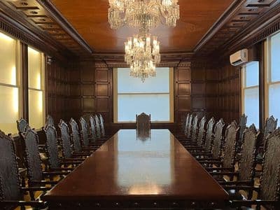 Large, elegant boardroom with a long, polished wooden table. Ornate chairs line both sides. Two chandeliers hang overhead, casting a warm glow.