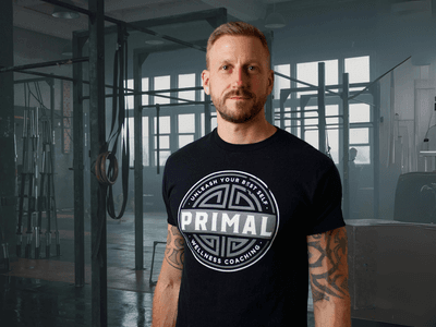 A man wearing a "PRIMAL Wellness Coaching" t-shirt stands in a gym, surrounded by fitness equipment, looking confidently at the camera.