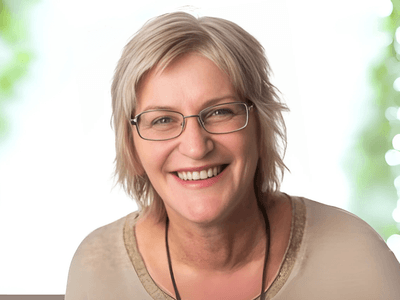 Smiling woman with short blonde hair and glasses, wearing a light-colored shirt, with a soft, blurred background of greenery and bokeh lights, radiating a warm and welcoming demeanor.
