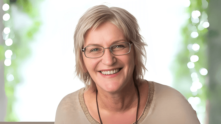 Smiling woman with short blonde hair and glasses, wearing a light-colored shirt, with a soft, blurred background of greenery and bokeh lights, radiating a warm and welcoming demeanor.