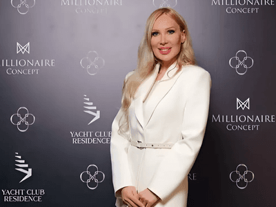 A woman in a white tailored blazer poses confidently in front of a branded backdrop featuring logos from 'Millionaire Concept' and 'Yacht Club Residence.' She smiles while standing with her hands at her sides, radiating elegance and professionalism.
