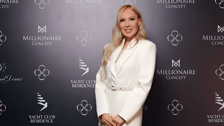 A woman in a white tailored blazer poses confidently in front of a branded backdrop featuring logos from 'Millionaire Concept' and 'Yacht Club Residence.' She smiles while standing with her hands at her sides, radiating elegance and professionalism.