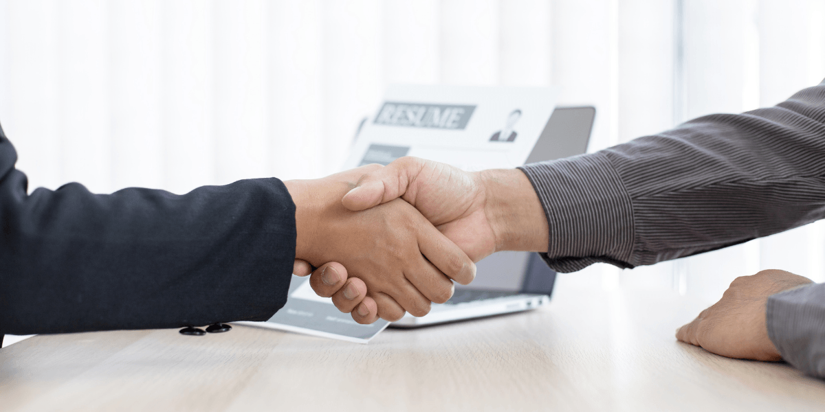 Two individuals shaking hands over a table with a laptop in the background