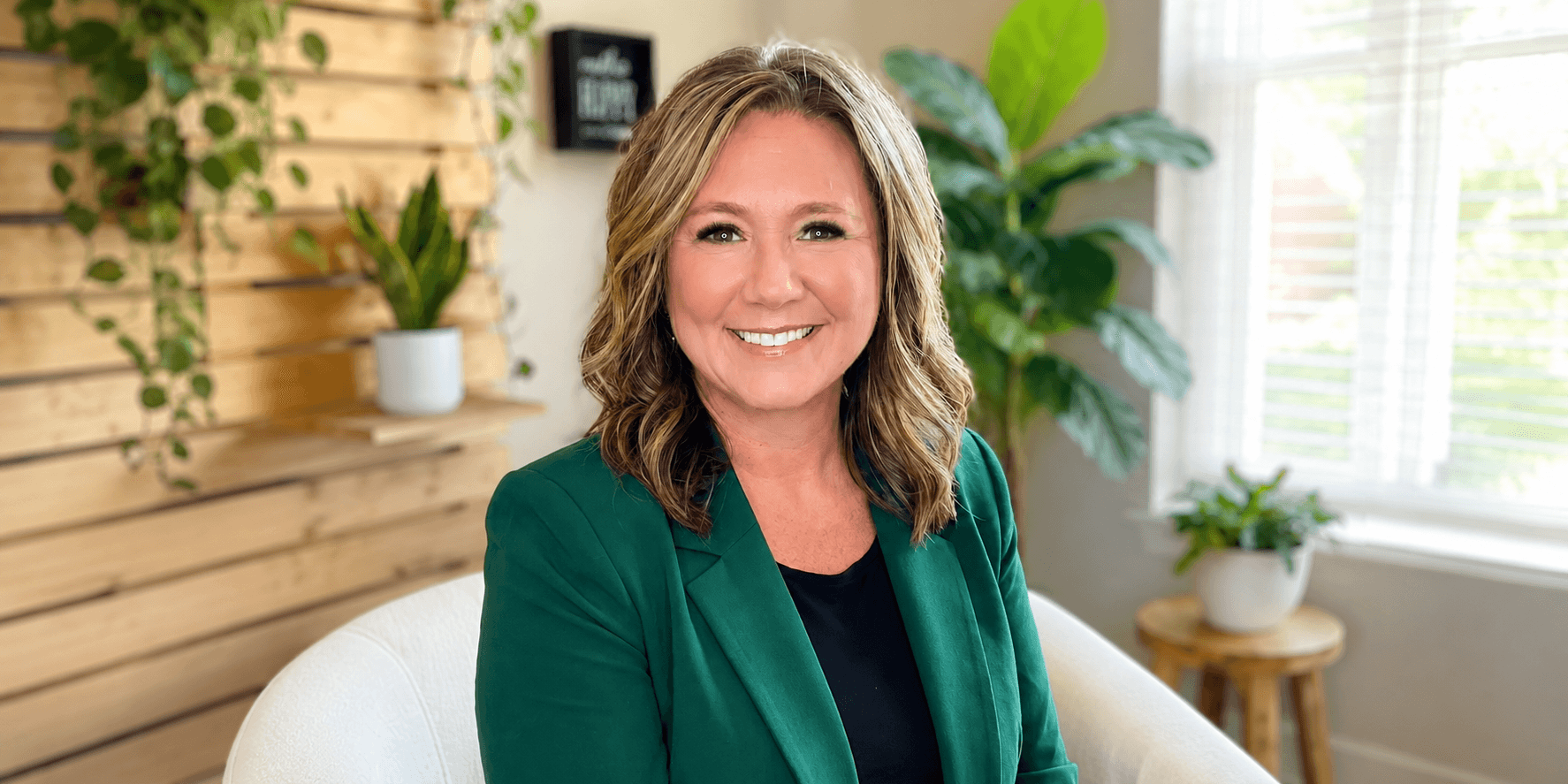 Smiling woman in a green blazer sits in a bright, plant-filled room