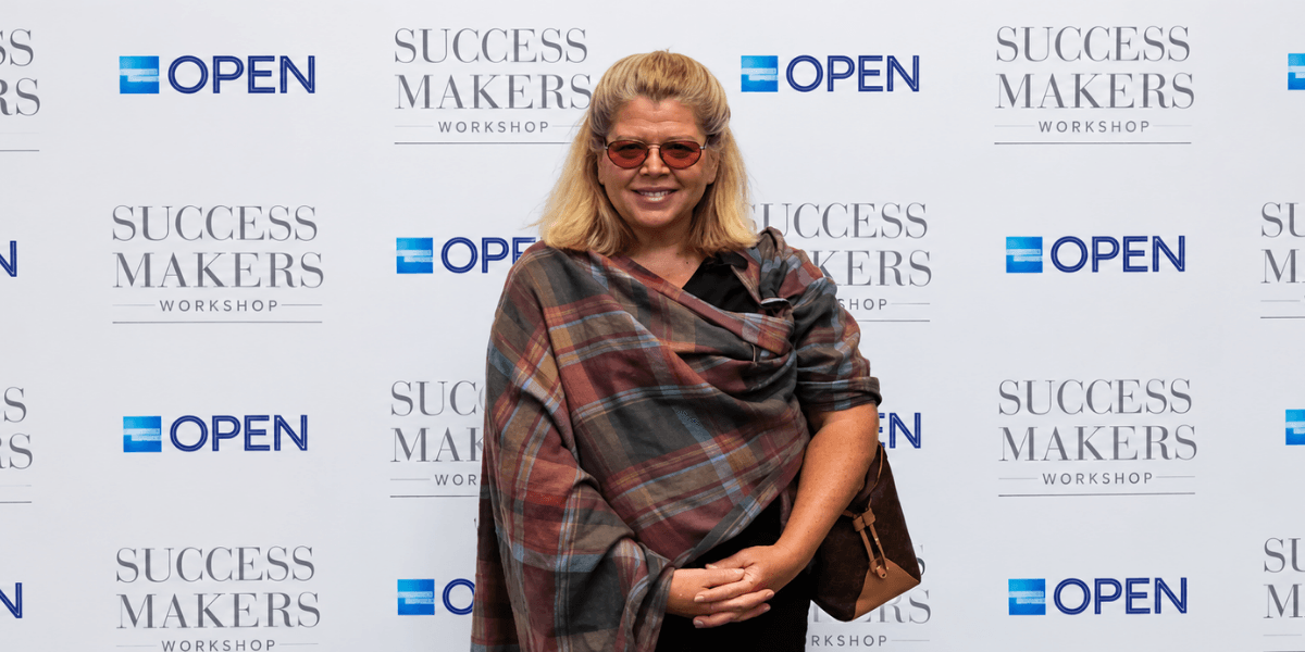 Smiling woman in sunglasses poses in front of a branded backdrop at an event