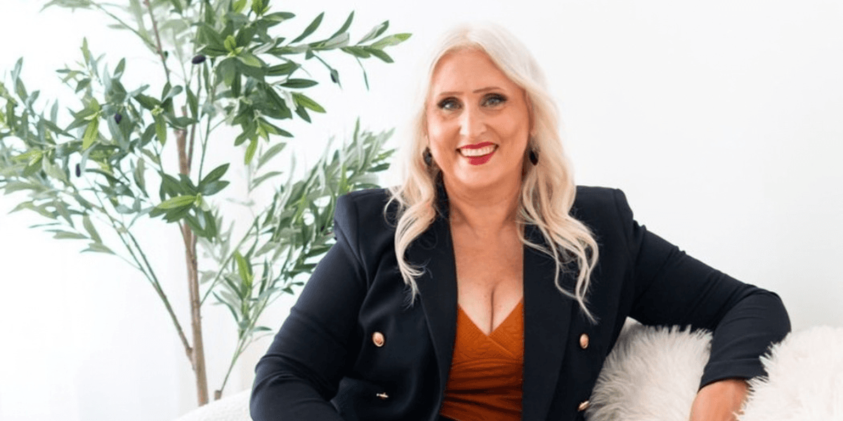 Smiling woman in a black blazer poses on a sofa near a plant