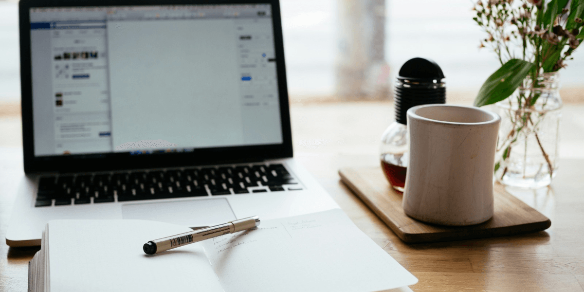 A laptop, notebook, pen, coffee cup, and flowers on a wooden table.