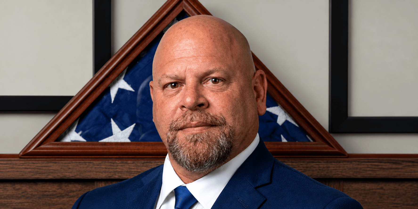 Man with a shaved head in a suit, standing in front of a flag display
