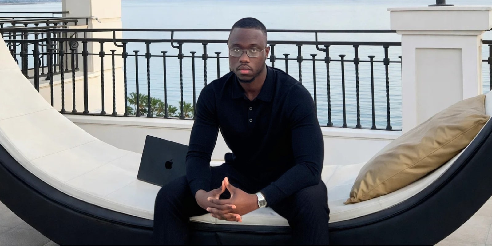 A man in a black polo shirt sits outdoors by the ocean with an Apple laptop, reflecting on sales systems.