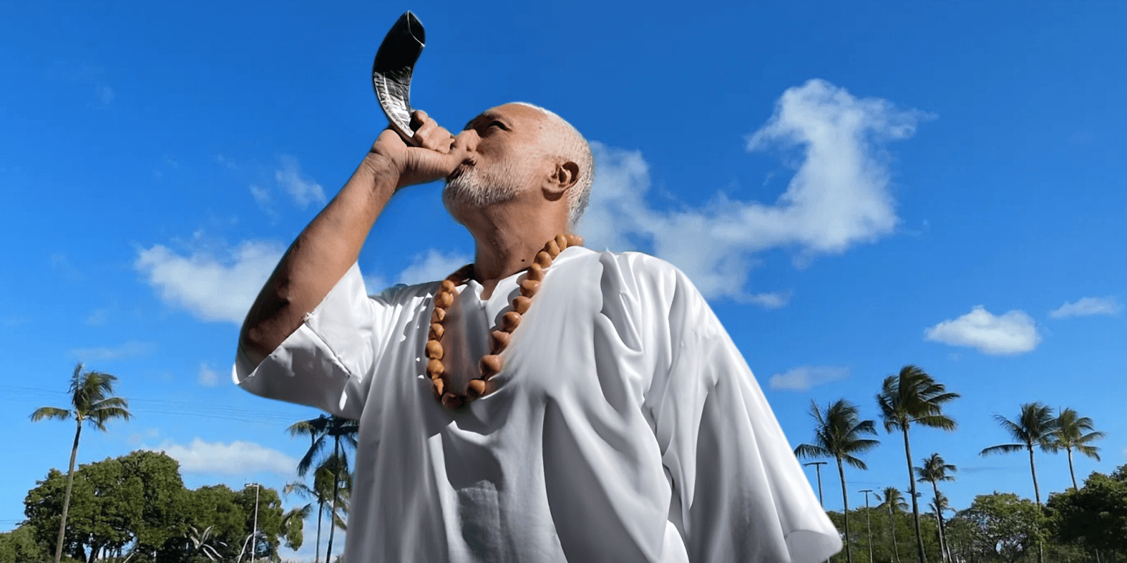 Man dressed in white, blowing a horn under a bright sky, symbolizing spiritual outreach and unity.