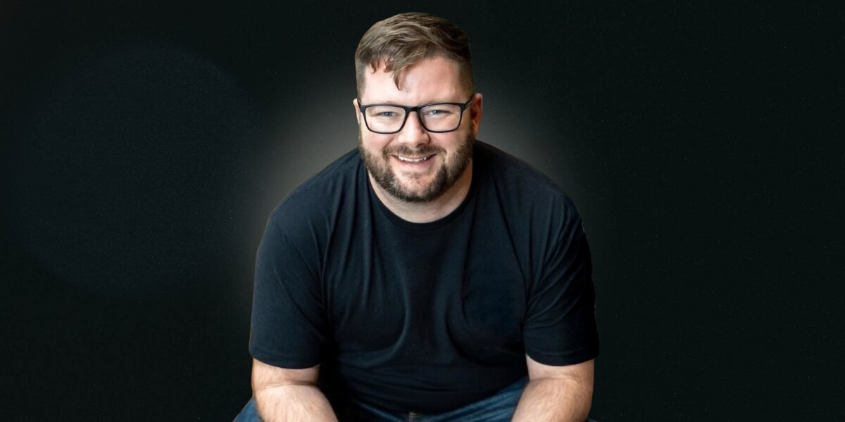 Will Craig smiling in glasses and a black shirt against a dark background.