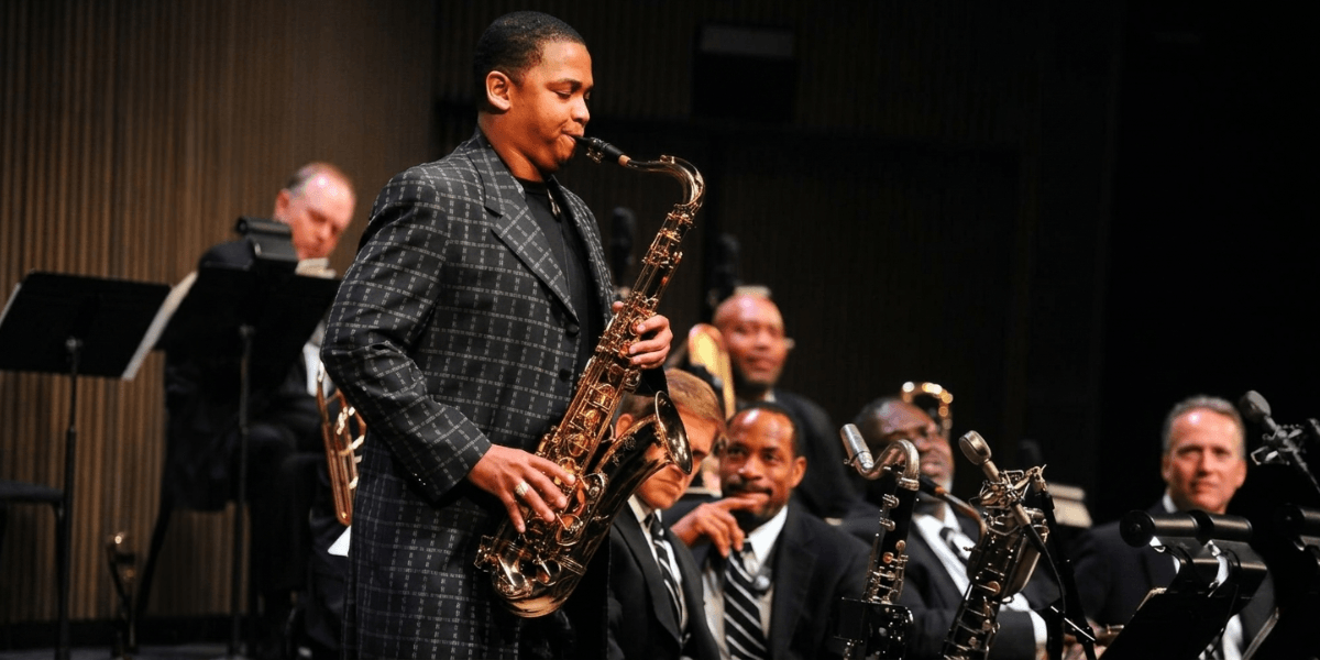 Jazz saxophonist performing solo on stage with orchestra seated behind him.