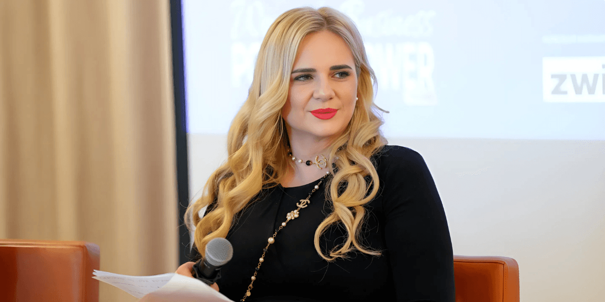 Blonde woman in a black dress with a Chanel necklace, holding a microphone and looking off to the side during a panel discussion.