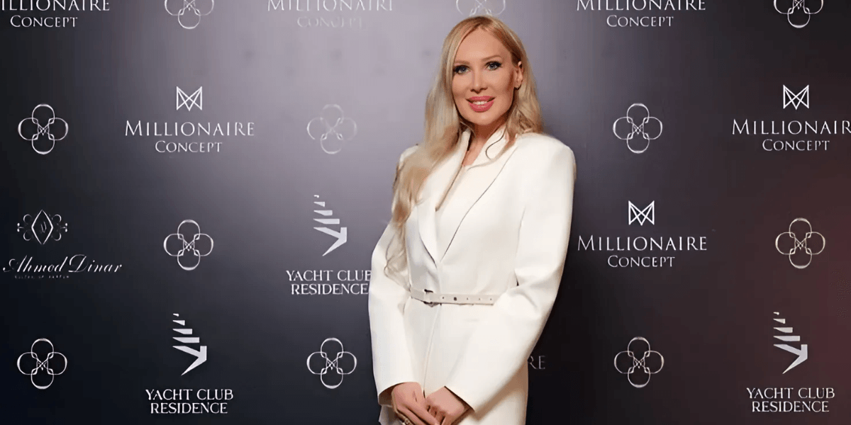 A woman in a white tailored blazer poses confidently in front of a branded backdrop featuring logos from 'Millionaire Concept' and 'Yacht Club Residence.' She smiles while standing with her hands at her sides, radiating elegance and professionalism.