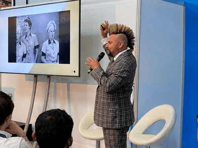 A presenter in a checked suit gestures towards a screen showing vintage images.