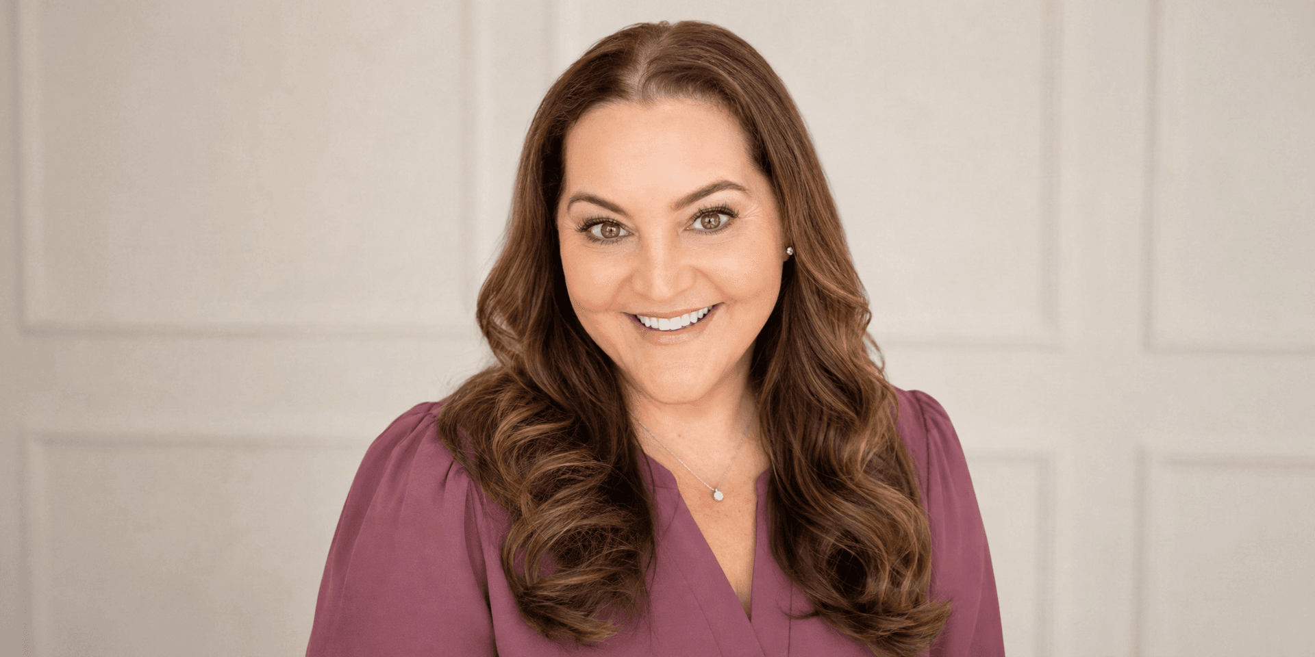 Smiling woman with wavy brown hair wearing a purple top against a neutral background
