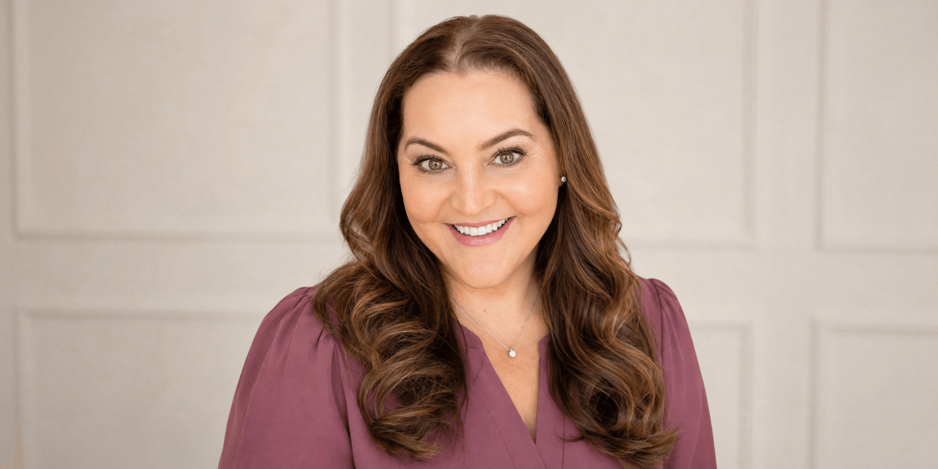 Smiling woman with wavy brown hair wearing a purple top against a neutral background