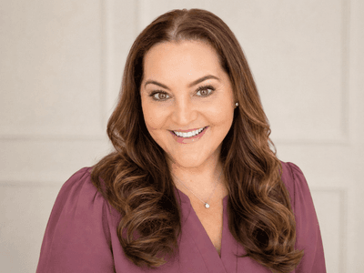 Smiling woman with wavy brown hair wearing a purple top against a neutral background