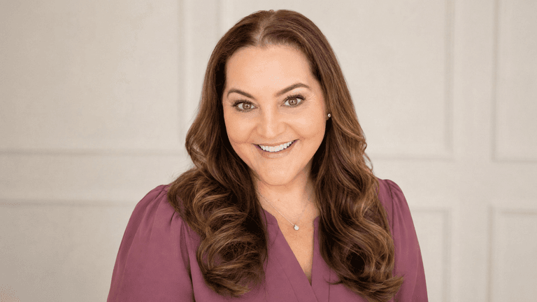 Smiling woman with wavy brown hair wearing a purple top against a neutral background