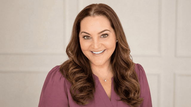 Smiling woman with wavy brown hair wearing a purple top against a neutral background