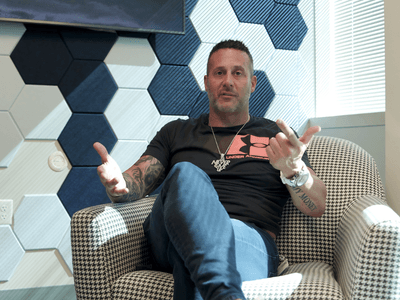 Man sitting in a chair, gesturing with hands, in a patterned room setting