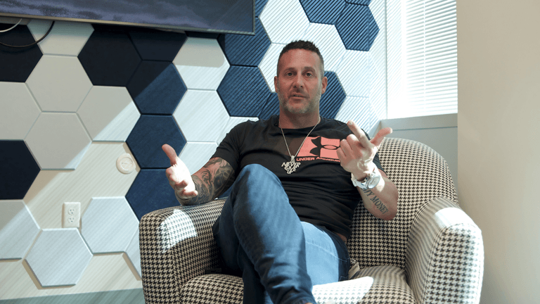 Man sitting in a chair, gesturing with hands, in a patterned room setting