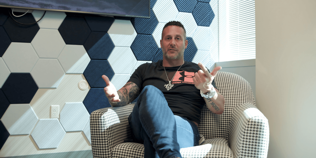 Man sitting in a chair, gesturing with hands, in a patterned room setting