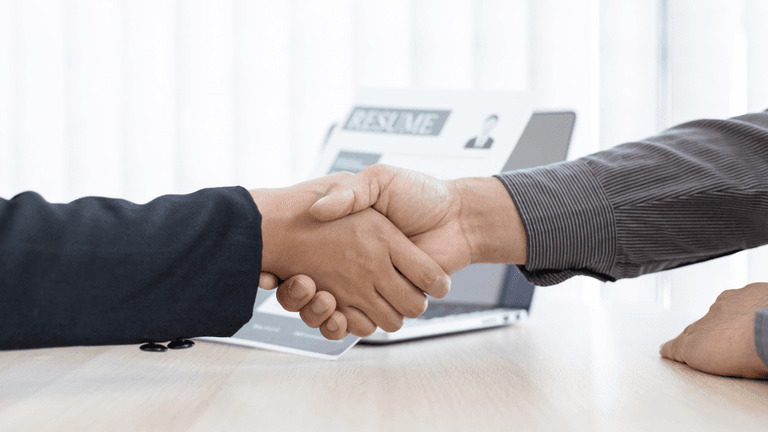 Two individuals shaking hands over a table with a laptop in the background