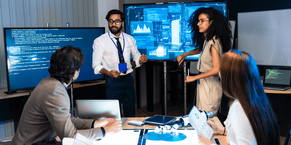 A diverse team collaborates around a table with screens displaying data and visuals.