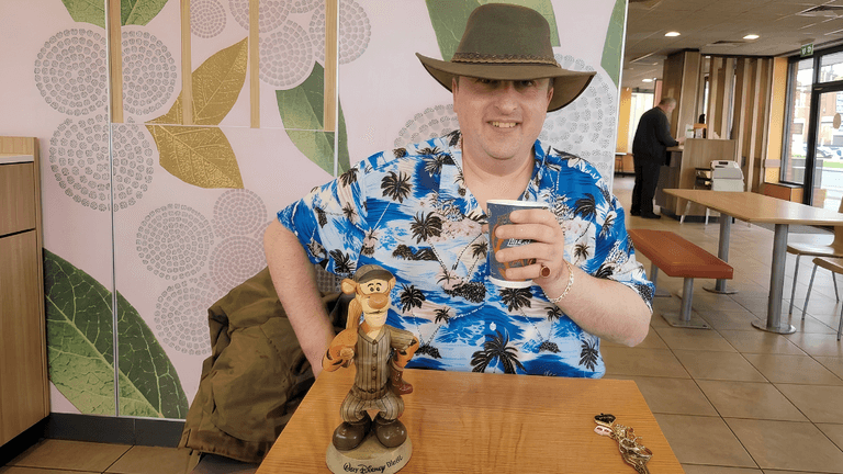 A man in a floral shirt and hat sits at a table with a toy.
