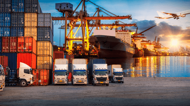 Shipping containers and trucks at a busy port with ships and an airplane in the background