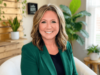 Smiling woman in a green blazer sits in a bright, plant-filled room