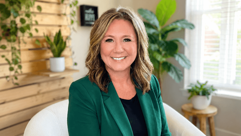 Smiling woman in a green blazer sits in a bright, plant-filled room