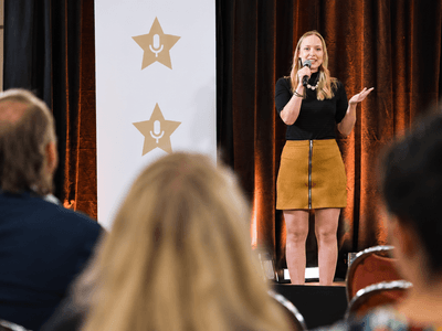 A woman stands on stage speaking to an audience at an event