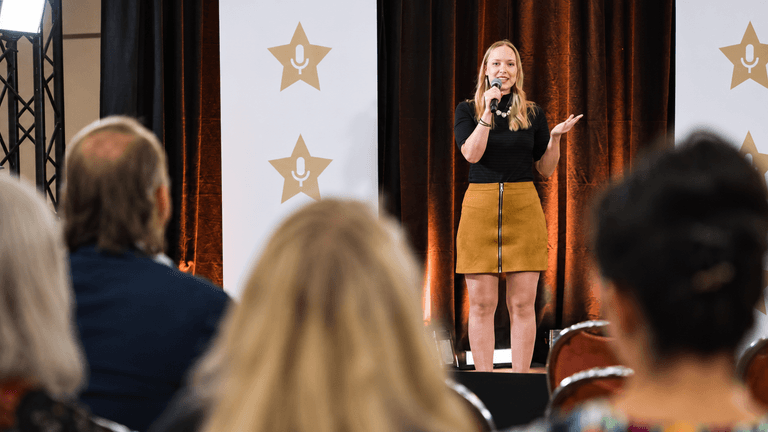 A woman stands on stage speaking to an audience at an event