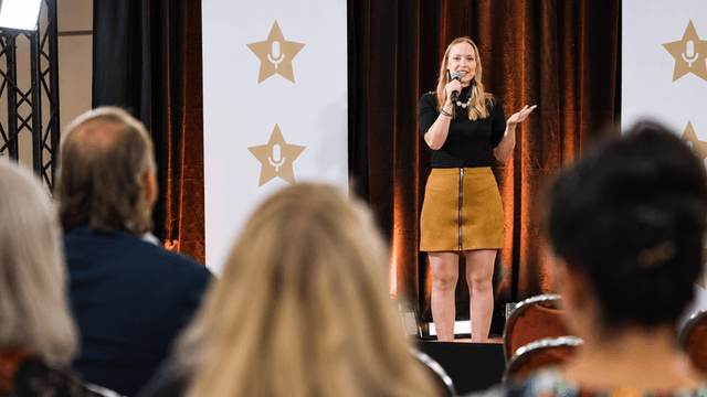 A woman stands on stage speaking to an audience at an event