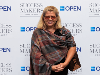Smiling woman in sunglasses poses in front of a branded backdrop at an event