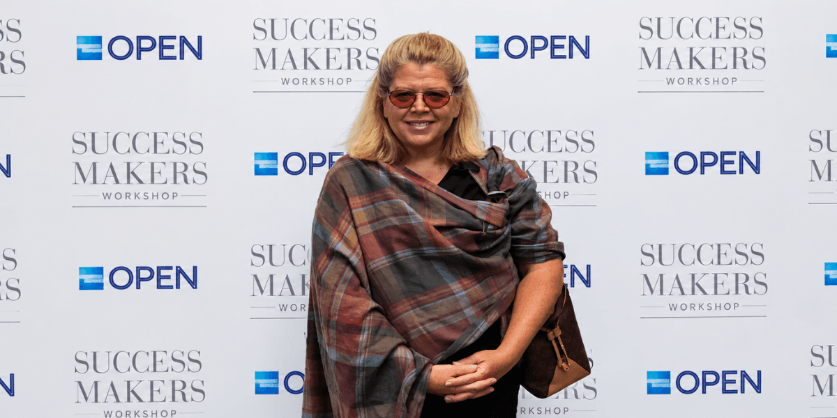 Smiling woman in sunglasses poses in front of a branded backdrop at an event