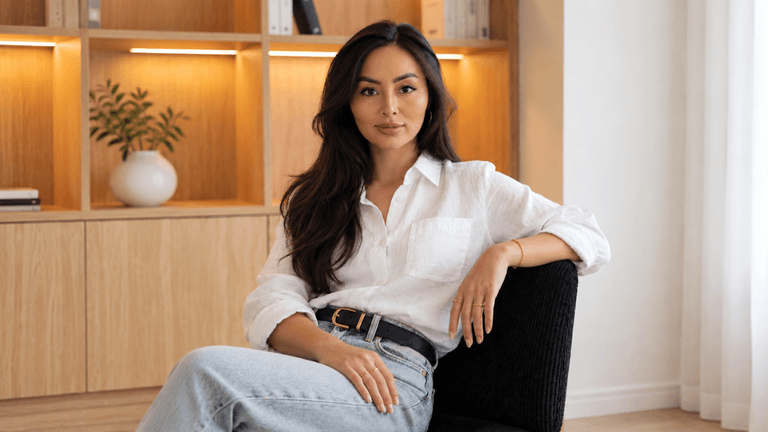 A woman with long dark hair sits confidently in a modern, minimalistic room