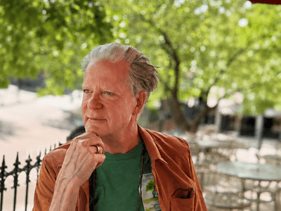 Man in a green shirt and brown jacket pondering outdoors with greenery in the background