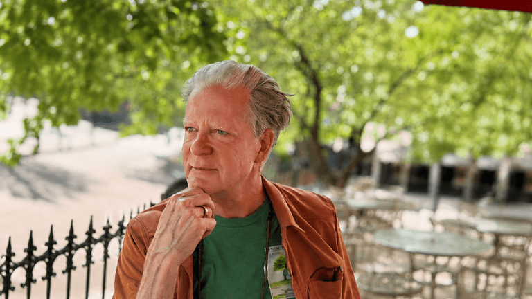Man in a green shirt and brown jacket pondering outdoors with greenery in the background