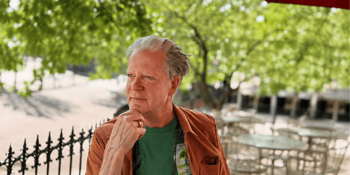 Man in a green shirt and brown jacket pondering outdoors with greenery in the background