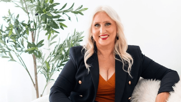 Smiling woman in a black blazer poses on a sofa near a plant